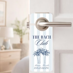 Op de afbeelding: Een deurhanger met een blauw en wit gestreepte achtergrond. De tekst "THE BACH Club" staat in elegant schrift boven een afbeelding van drie palmbomen. Daaronder staat de tekst "Miami, Florida ANNA'S ROOM".