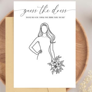Pode incluir: Um cartão branco com o texto "adivinhe o vestido" e um desenho de uma mulher em um vestido segurando flores. O cartão está em um prato de madeira com um envelope bege.