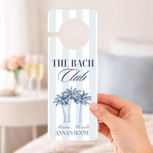 May include: A light blue and white striped door hanger with the text "THE BACH Club" and a palm tree illustration. The text "Miami, Florida ANNA'S ROOM" is also printed on the door hanger.