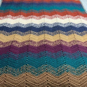 May include: A handmade, colorful crochet blanket with a chevron pattern. The blanket features a variety of colors, including brown, teal, purple, gold, and cream. The blanket is made of soft yarn and is perfect for snuggling up on the couch or bed.