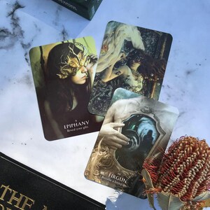 Może przedstawiać: Trzy karty tarota z talii "The Art of Witch", przedstawiające ilustracje kobiety z maską z liści, kobiety leżącej na łożu z liści i dłoni trzymającej lustro. Karty są oznaczone jako "Epiphany", "Reveal your gifts", "Firgun" i "Become a loving mirror".