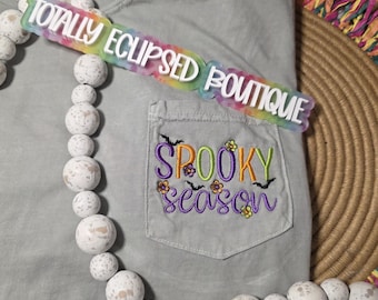 May include: A light gray t-shirt with a pocket embroidered with the words "Spooky Season" in purple, green, and yellow. A rainbow-colored sign that says "Totally Eclipsed Boutique" is on top of the shirt. A necklace with white speckled beads is in front of the shirt.