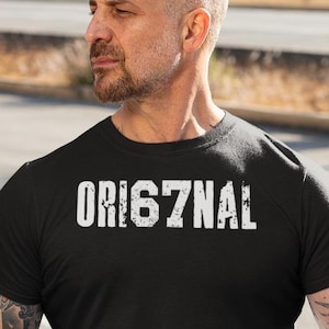May include: Black t-shirt with the word "ORIG7NAL" in white lettering. The person is wearing blue jeans and is sitting on a black motorcycle. A black helmet with a checkered pattern is visible.