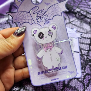 May include: A purple phone grip featuring a white teddy bear with a stitched design, a cross-shaped eye, and a spiral eye. The grip is on a purple backing with the text "FRAFRANE GRIP" and "MUHAN ARI".