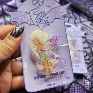 May include: A phone grip featuring a fairy illustration with long white hair and butterfly wings, mounted on a light purple backing card. The card displays the text Frafran Art, Frafran Phone Grip, and Human Art, decorated with bat and spiderweb graphics.