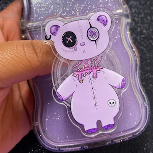 May include: A clear phone case with a purple glitter background, holding a purple teddy bear sticker. The sticker has a stitched design, a skull, and a button eye. The bear's neck is open with pink tendrils.