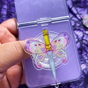 May include: A clear phone grip featuring a butterfly design with a dagger. The butterfly wings are decorated with hearts and swirls in pink, purple, and white. The dagger has a yellow handle and a light blue blade. The grip is attached to a purple phone case.