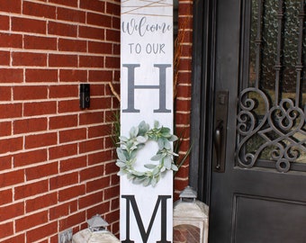 Può includere: Un cartello di legno bianco con la scritta "Welcome to our Home" in lettere nere. Il cartello è decorato con una ghirlanda verde e spago. Il cartello è appoggiato a un muro di mattoni accanto a una porta nera con un tappetino di benvenuto che dice "hello".