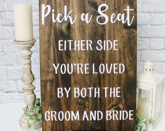 May include: A rustic wooden sign with white text that reads "Pick a Seat, Either Side, You're Loved By Both The Groom and Bride".