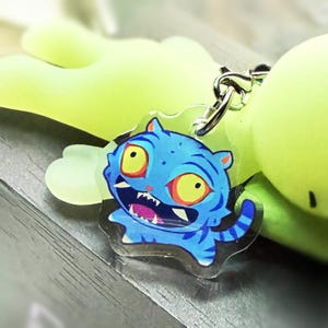 Peut inclure: Un porte-clés en forme de tigre de dessin animé bleu et blanc avec un anneau en métal argenté. Le tigre a des yeux jaunes et un large sourire. Le porte-clés est attaché à un objet vert clair.