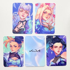 May include: Set of five rectangular K/DA fan art cards. The cards feature illustrations of female characters in vibrant colors, with the K/DA logo on the back. The artwork includes shades of blue, purple, pink, and gold.