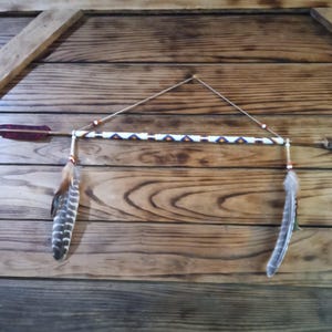 May include: A decorative arrow and feather wall hanging. The arrow has a red fletching and is adorned with a beaded section in white, blue, and orange. Two feathers hang below the arrow, and the background is a wooden surface.