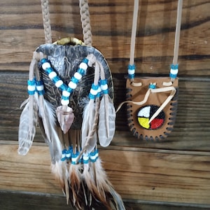 May include: Two handcrafted Native American-style necklaces. One features a turtle shell pendant adorned with blue and white beads, feathers, and a stone arrowhead. The second is a small leather pouch with a beaded design and leather cord.