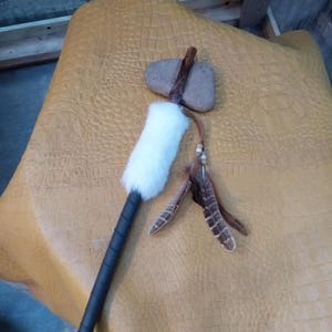 May include: A brown wooden handle with a gray stone head, wrapped in white fur and decorated with feathers. The handle is wrapped in black leather and has a leather strap at the bottom. The stone head is shaped like a hammer.