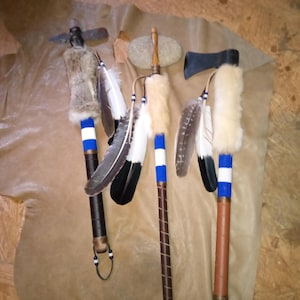 May include: Four ceremonial staffs with blue and white paint, feathers, and fur accents. The staffs have different heads, including a stone, a wooden axe, and a wooden pipe.