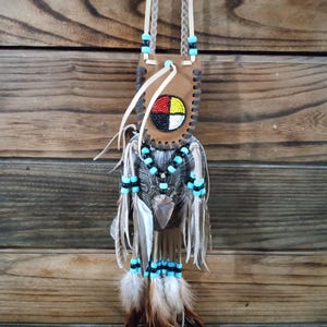 May include: A Native American-style pendant with a leather pouch featuring a colorful circular design. The pendant is adorned with turquoise beads, feathers, and a braided leather strap. The feathers are brown and white.
