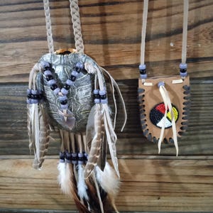 May include: Two handcrafted Native American necklaces. One features a turtle shell pendant adorned with beads and feathers. The other is a small leather pouch with beaded details and fringe. Both hang against a wooden background.