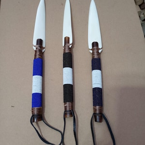 May include: Three decorative spears featuring white pointed blades and wooden shafts wrapped in colorful seed beads. Each handle displays a unique pattern of blue, white, black, or dark purple beads and includes a black leather wrist loop at the base.