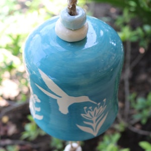 May include: A blue ceramic wind chime with a white hummingbird and flower design. The chime is hanging from a string with three white and blue ceramic beads.