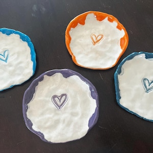 May include: Four handmade ceramic dishes with irregular edges. Each dish has a white interior and a different colored rim: blue, orange, purple, and teal. A heart is painted in the center of each dish.