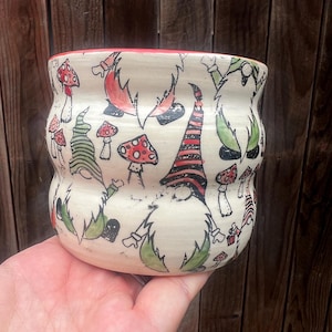 May include: A ceramic mug with a cream-colored base featuring a repeating pattern of gnomes with long beards and pointed hats in shades of green, red, and black, interspersed with illustrations of red and white spotted mushrooms and a red rim.