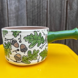 May include: A ceramic soup mug with a cream-colored base featuring a hand-painted pattern of green oak leaves and brown acorns. The mug has a smooth, glossy finish and a solid green handle extending from the side, with a light pink interior.