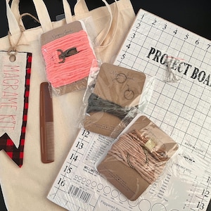 May include: A macrame kit with a white canvas tote bag, a wooden comb, and four spools of cord. The kit includes a project board with a grid pattern and the words "Project Board" printed on it. The tote bag has a red and black plaid tag with the words "Macrame Kit" printed on it.