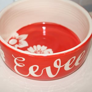 May include: Red ceramic pet bowl with white floral designs and the name "Evel" written in white cursive.