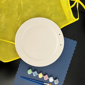 May include: A white ceramic plate with a scalloped edge and a blue paint mat with paint pots and brushes. The plate is on top of a yellow smock.