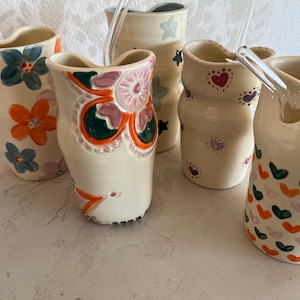 May include: Five ceramic tumblers with wavy rims and hand-painted designs. One features blue and orange flowers, another a large pink and orange floral motif, one has stars, one has small hearts, and the last has a pattern of orange and green hearts.