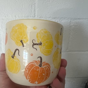 May include: A cream-colored ceramic mug featuring a repeating pattern of yellow and orange pumpkins with dark brown stems and vines. The pumpkins are illustrated in a simple, graphic style distributed evenly across the exterior surface of the mug.