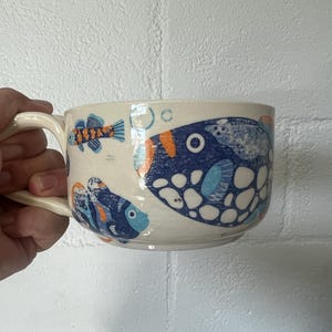 May include: A wide, cream-colored ceramic mug featuring a hand-painted pattern of various blue and orange fish, including a large pufferfish, smaller tropical fish, and light blue bubbles scattered across the exterior surface with a curved handle.