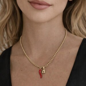 Puede incluir: Un collar de cuentas doradas con dos colgantes. Uno es un pimiento rojo, y el otro es una letra "A" dorada. El collar se lleva sobre una camiseta negra.