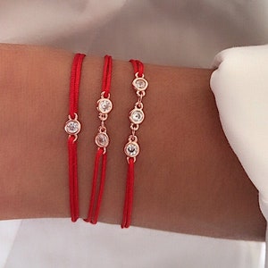 Puede incluir: Tres pulseras de cuerda roja con pequeñas piedras preciosas transparentes en una cadena dorada. Las pulseras se llevan en la muñeca.