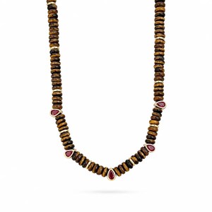 Puede incluir: Un collar que presenta una hilera de cuentas cilíndricas con rayas marrones y negras, intercaladas con pequeños espaciadores de tono dorado. El diseño incluye cuatro piedras preciosas rojas en forma de lágrima engastadas en metal dorado, dispuestas simétricamente en la parte inferior.