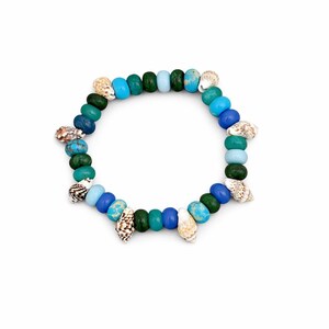 Puede incluir: Una pulsera de cuentas con cuentas azules, verdes y turquesas y pequeñas conchas marinas. La pulsera es circular sobre un fondo blanco. Las cuentas tienen aproximadamente 1,3 cm de diámetro.