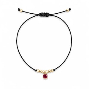 Puede incluir: Una pulsera de cordón negro con un dije central de piedra roja de corte cuadrado, flanqueado por cuatro pequeñas cuentas esféricas doradas a cada lado. El cierre ajustable incluye un mecanismo de nudo corredizo y dos cuentas doradas en los extremos.