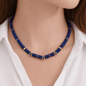 Puede incluir: Un collar de cuentas azul oscuro con detalles dorados y colgantes en forma de lágrima. El collar se lleva sobre una camisa blanca.