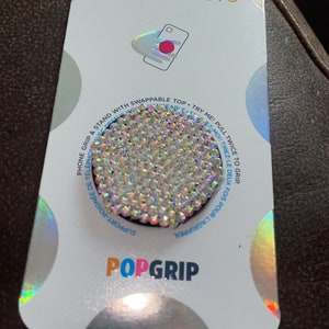 Könnte beinhalten: Eine weiße Karte mit holografischem Finish, auf der ein PopSockets PopGrip abgebildet ist. Der PopGrip ist ein runder, transparenter und glitzernder Handygriff mit schwarzer Basis. Auf der Karte steht der Text "POPSOCKETS" und "POPGRIP" gedruckt. Die Karte enthält auch Anweisungen zur Verwendung des PopGrip.