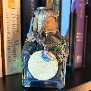 May include: A decorative glass bottle filled with iridescent blue and green fragments. The bottle has a silver wax seal and a charm with a moon and star. The label reads "Dreams and Wishes."