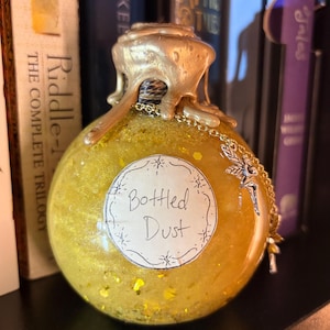 May include: A round, yellow glass bottle filled with glitter and liquid, labeled "Bottled Dust." The bottle has a gold-colored, dripped wax seal and a decorative chain with a fairy charm. The bottle is displayed in front of a bookshelf.