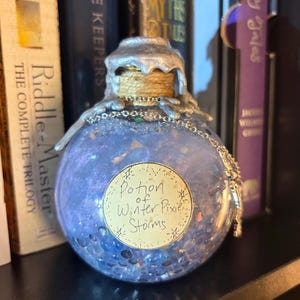 May include: A round, blue potion bottle with a silver-colored top and a label that reads "Potion of Winter Pixie Storms." The bottle is filled with glitter and small, round objects. A silver chain hangs from the top of the bottle.