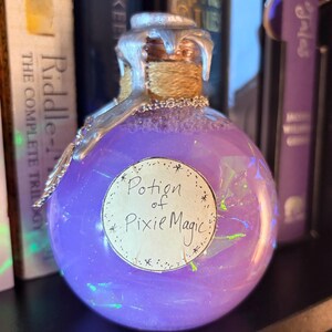 May include: A round glass bottle filled with a shimmering purple liquid, labeled "Potion of Pixie Magic." The bottle has a silver-colored top and a cork sealed with twine. The background includes books on a shelf.