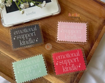 May include: Four rectangular stickers with the text "emotional support kindle" in white, each with a different colored background: dark gray, pink, mint green, and red. A Kindle e-reader and flowers are also in the image.
