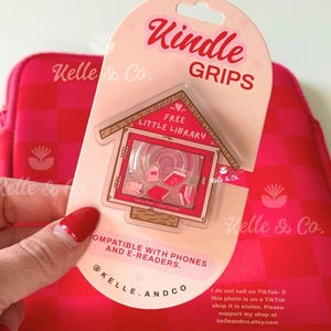 May include: A pink and red Kindle Grips package featuring a "Free Little Library" design. The package is held in front of a pink and red checkered case. Text on the package reads "Kindle Grips" and "Compatible with Phones and E-Readers."