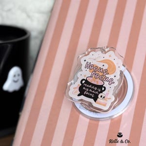 May include: A clear acrylic keychain with a Halloween theme. The keychain features the words "Hocus Pocus" and "reading is my main focus" with a cauldron, moon, and stars design. The keychain is on a pink and white striped background.