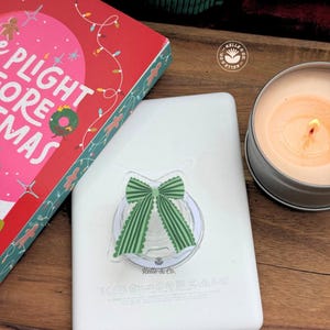 May include: A white phone grip featuring a green and white striped bow design with scalloped edges, attached to the back of a white electronic device. The device is positioned next to a red book titled The Plight Before Christmas and a lit candle.