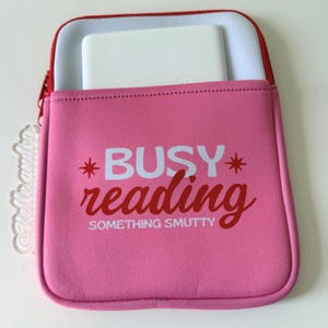 May include: Pink neoprene tablet sleeve with a red zipper and trim. The sleeve features the text "BUSY reading SOMETHING SMUTTY" in white and red. A white tablet is partially visible inside the sleeve.