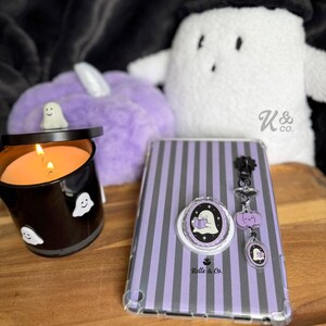 May include: A Halloween-themed collection featuring a black candle with ghost decals, a purple striped tablet case, and a matching pop socket. A plush purple pumpkin and a white ghost plush are also visible.