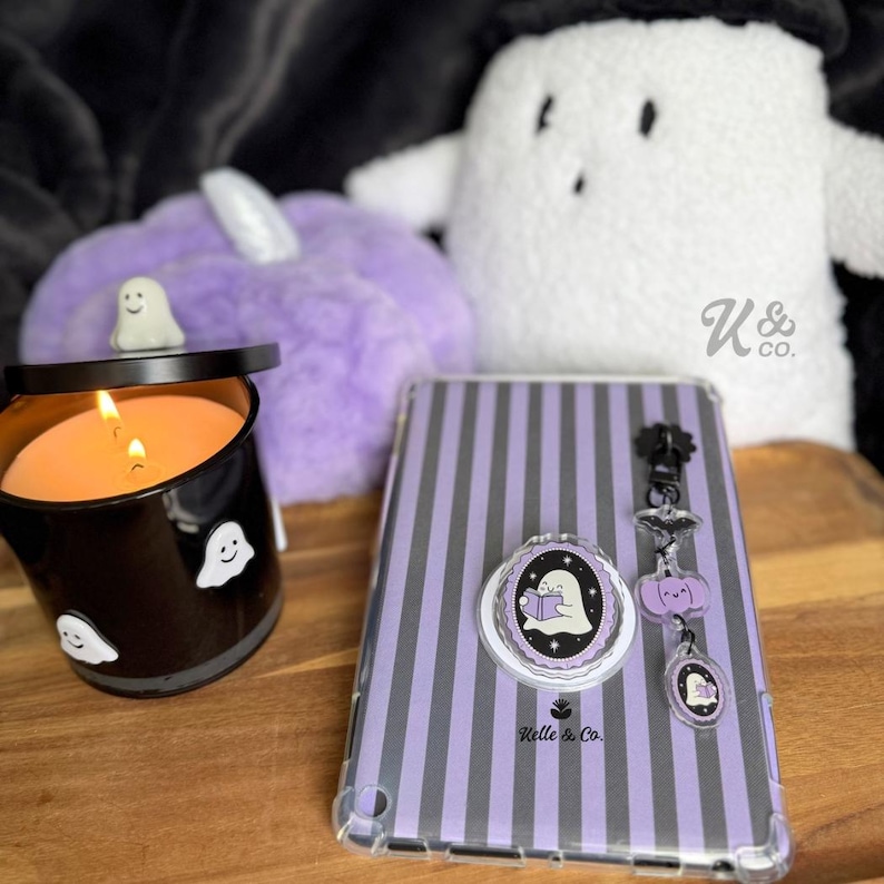 May include: A Halloween-themed collection featuring a black candle with ghost decals, a purple striped tablet case, and a matching pop socket. A plush purple pumpkin and a white ghost plush are also visible.